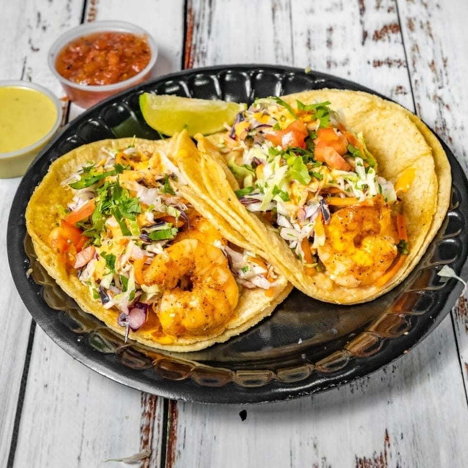 Two Bandeño Mexican Food shrimp tacos with cabbage, tomatoes, and cilantro, served with lime and sauces.