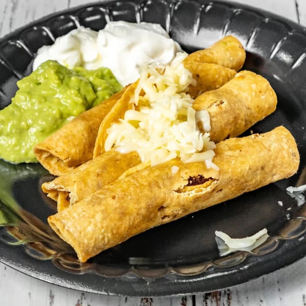Crispy taquitos topped with shredded cheese, served with guacamole and sour cream on a black plate.